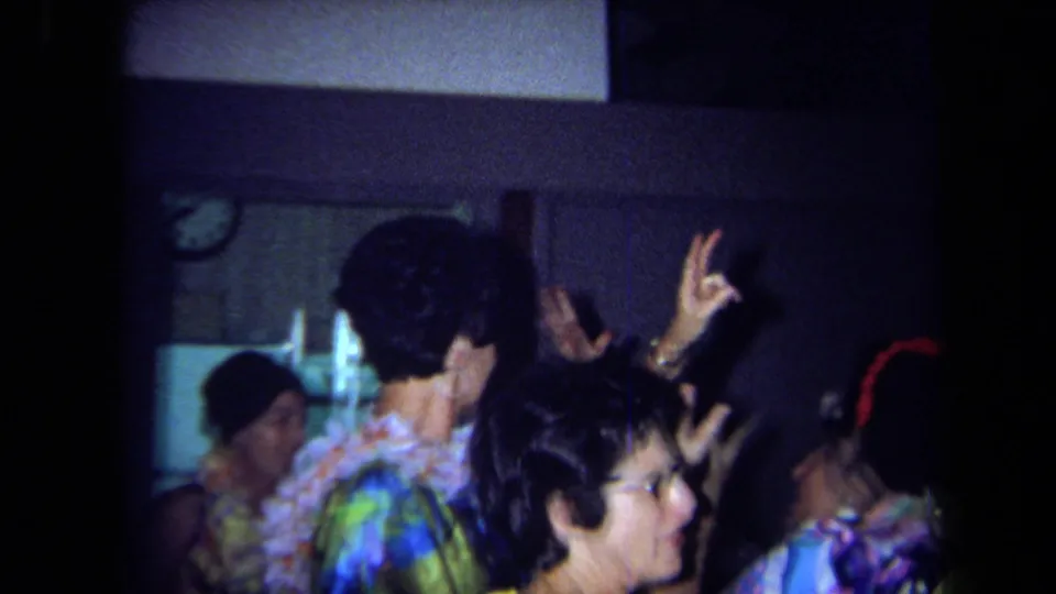 Adults wearing leis and colorful tropical clothing gather closely on a dim dance floor during the 1971 California reunion.