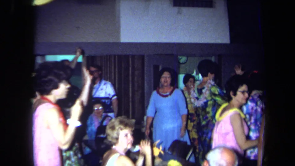 1971 California reunion footage shows seated and standing adults in leis and bright clothing dancing near curtains and a wall clock.