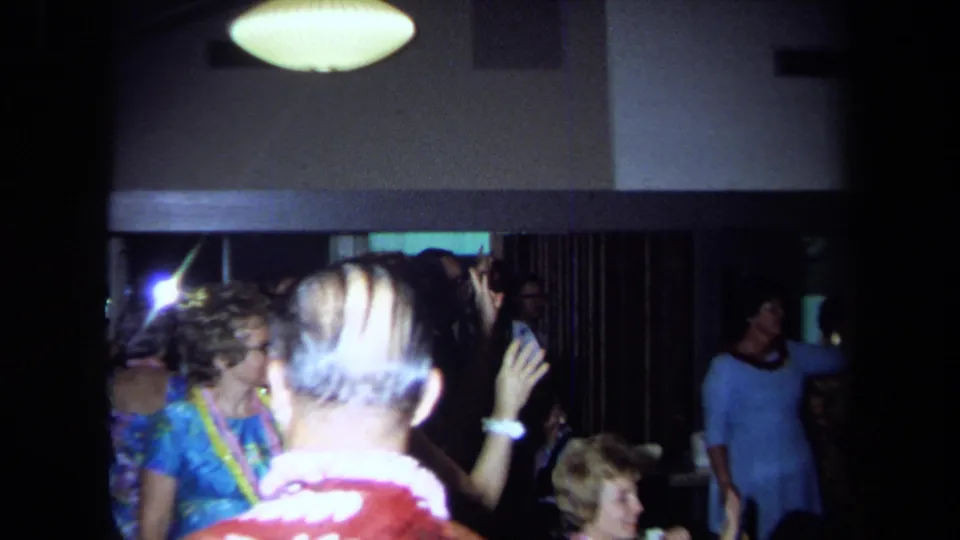 A 1971 California home-movie frame captures reunion guests in leis dancing beneath a ceiling light inside a private hall.
