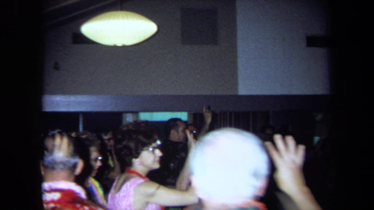 1971 California home-movie frame showing party guests in leis gathered in a dim interior beneath a pendant lamp.