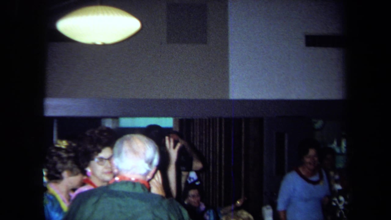 Party guests in leis crowd together under a ceiling lamp.