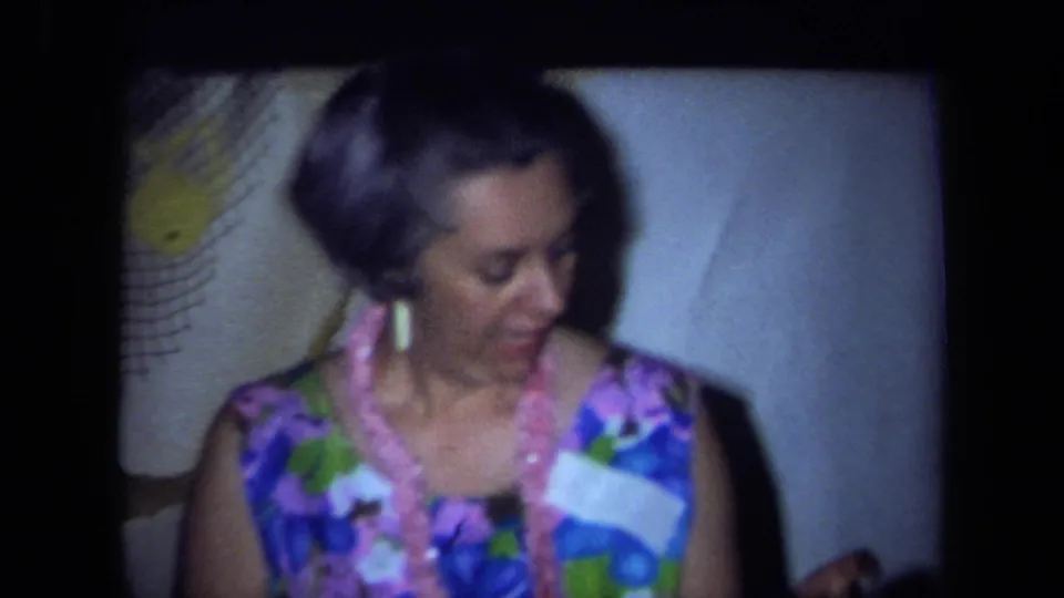 1971 California home-movie frame showing a woman in a floral dress and pink lei looking down during the party.