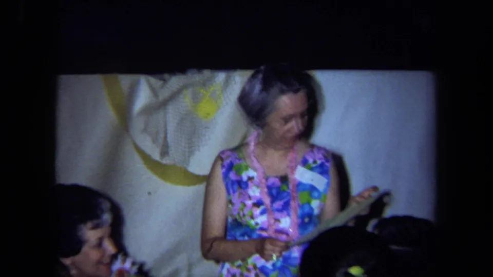 1971 California home-movie frame of a woman in floral clothing and lei appearing to read from a paper at the party.
