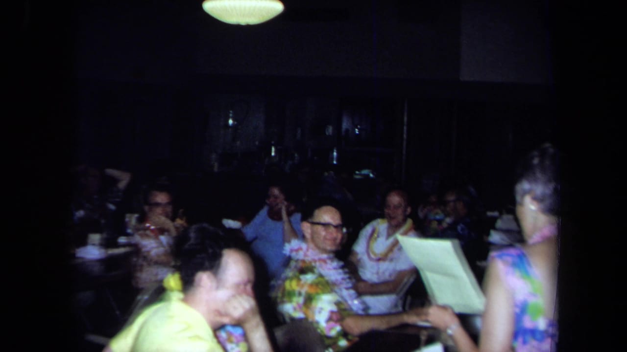 1971 California 8mm frame of adults at tables during an indoor gathering as a woman appears to read from paper.