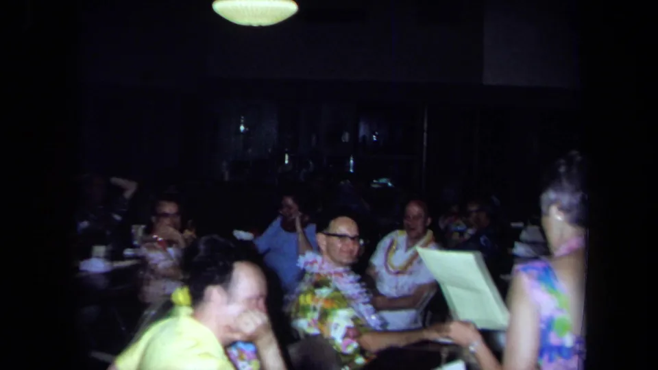 1971 California 8mm frame of adults at tables during an indoor gathering as a woman appears to read from paper.