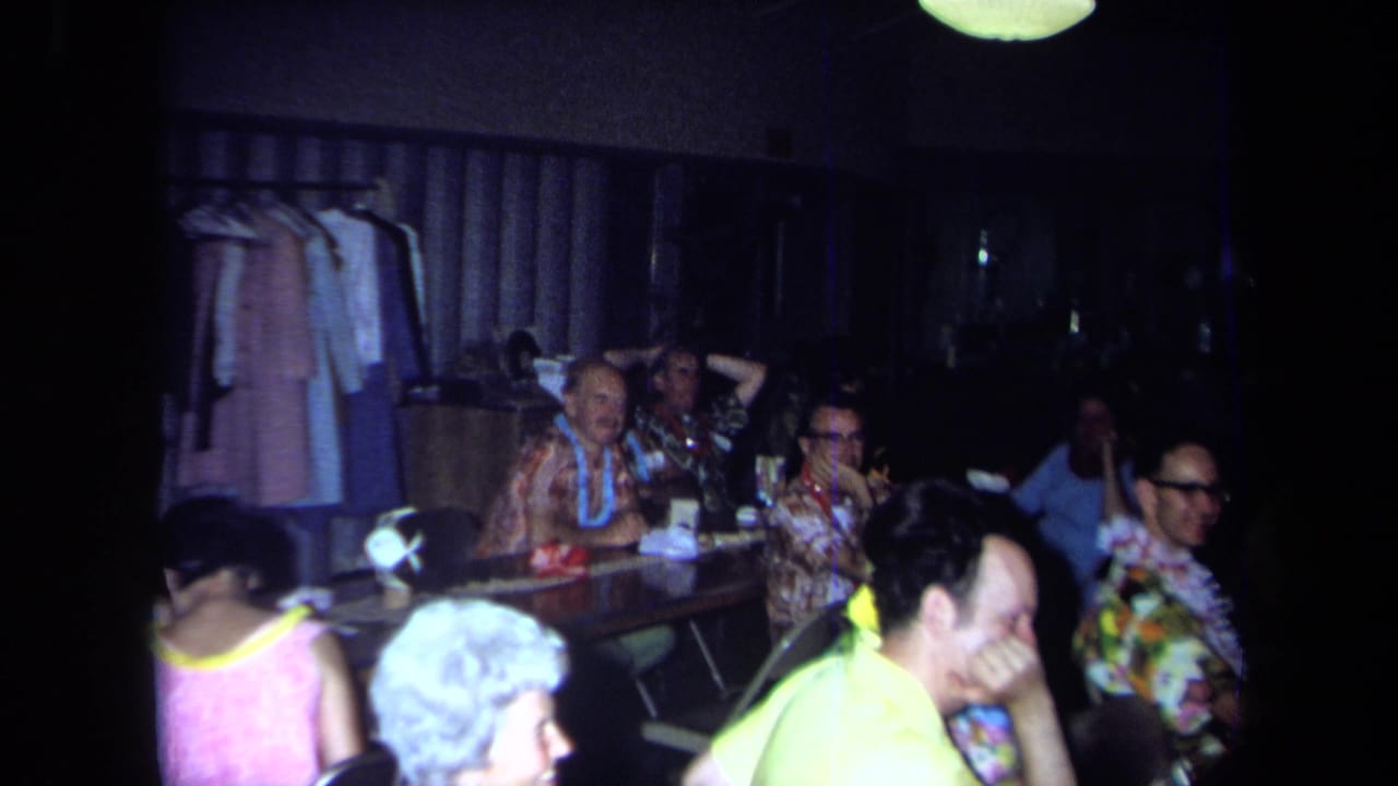 Adults sit at a long table with leis and floral clothing in a dim private room.