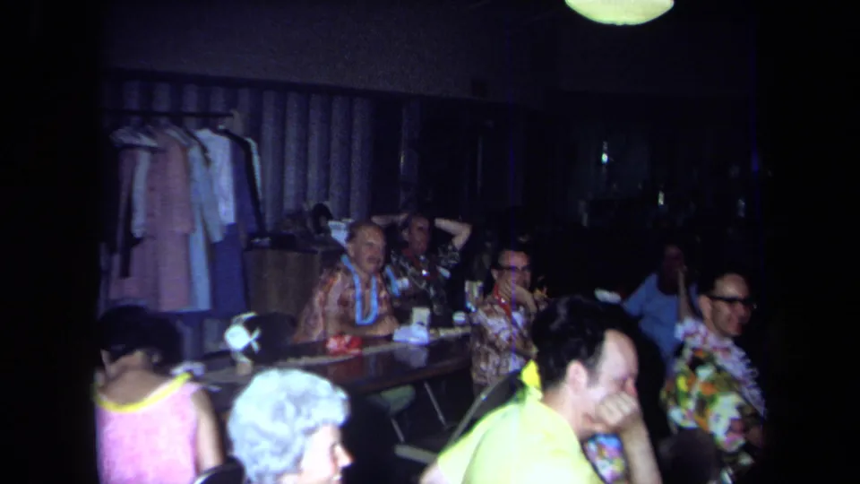 1971 California home-movie frame showing adults seated at a long table with leis, floral shirts, and a clothing rack behind them.