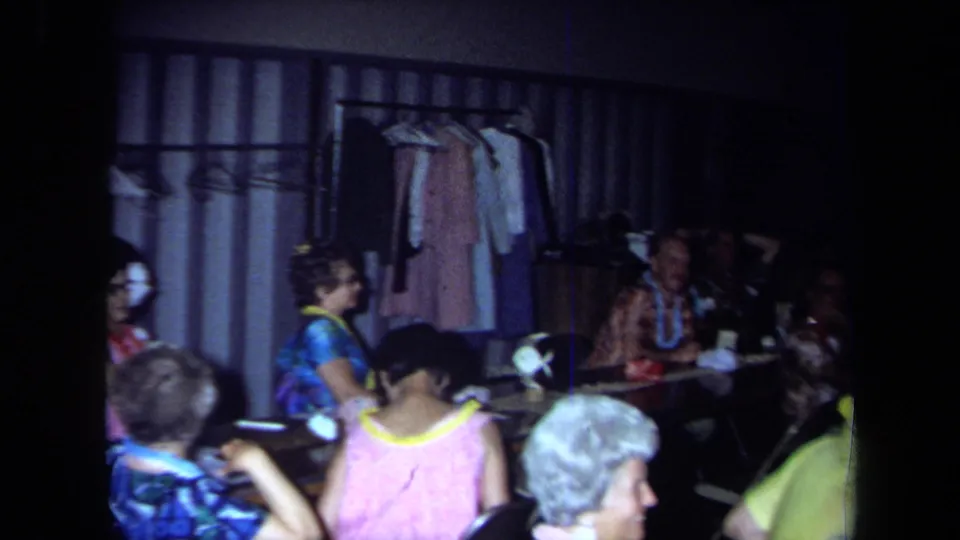 1971 California 8mm still of adults at an indoor party, with leis, patterned clothing, tableware, and coats on a rack in the background.