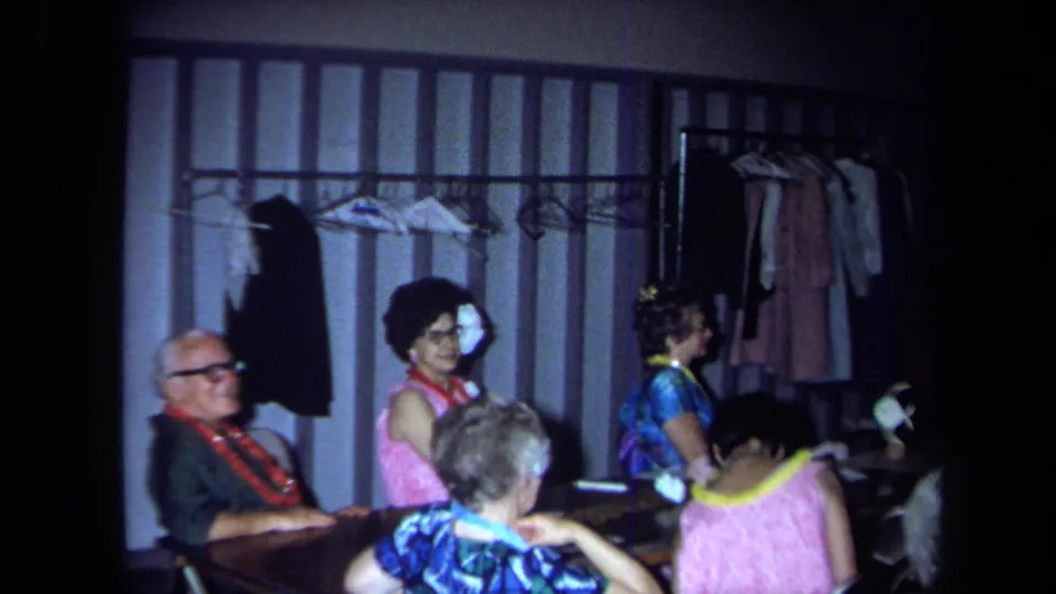 1971 California home-movie frame of adults seated by tables near a wall-mounted clothing rack during an indoor social gathering.