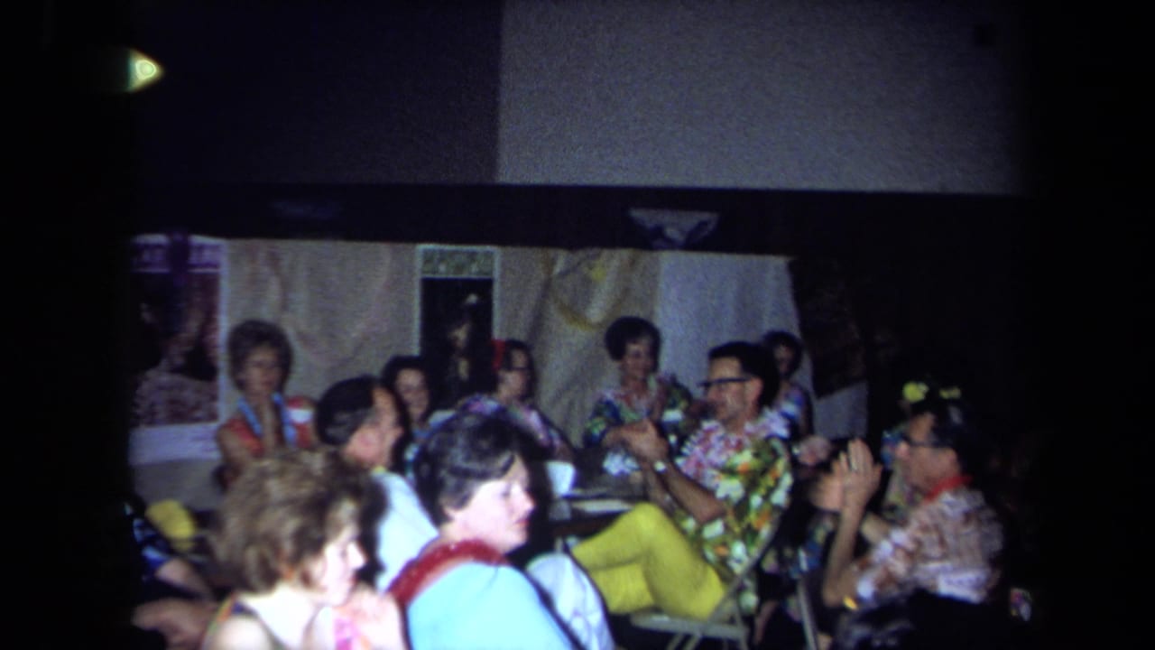 1971 California home-movie frame of older adults wearing leis and patterned shirts at a Hawaiian-themed indoor party.