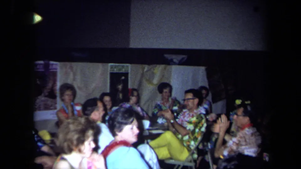 1971 California home-movie frame of older adults wearing leis and patterned shirts at a Hawaiian-themed indoor party.