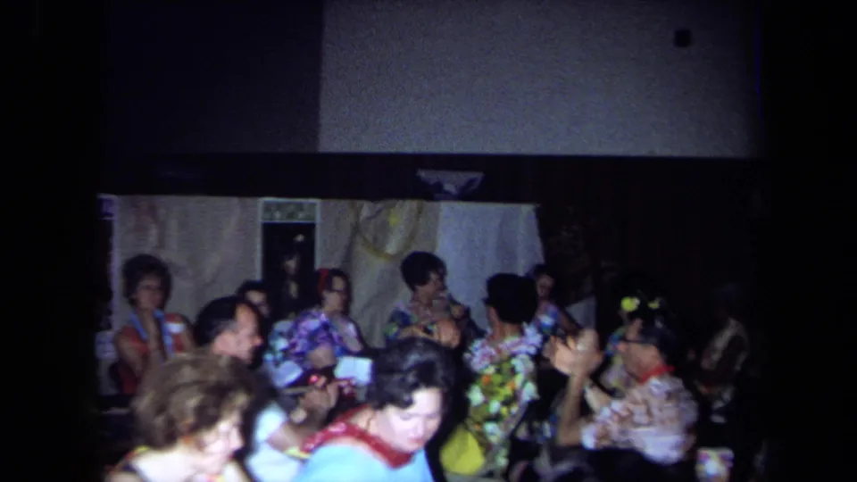 Older adults at a 1971 California Hawaiian-themed party sit together, with several guests clapping in the dim room.