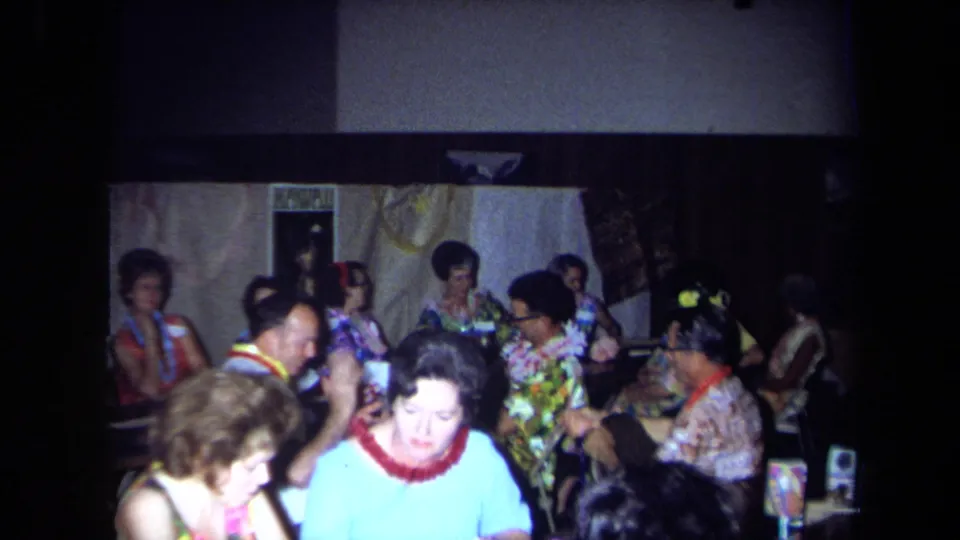 Crowded 1971 California home-movie view of older adults in leis and colorful clothing seated around party tables.