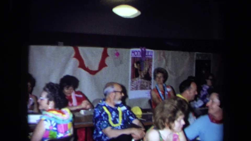 1971 California frame with older adults seated under Hawaiian-themed decor, including leis, posters, and wall garlands.
