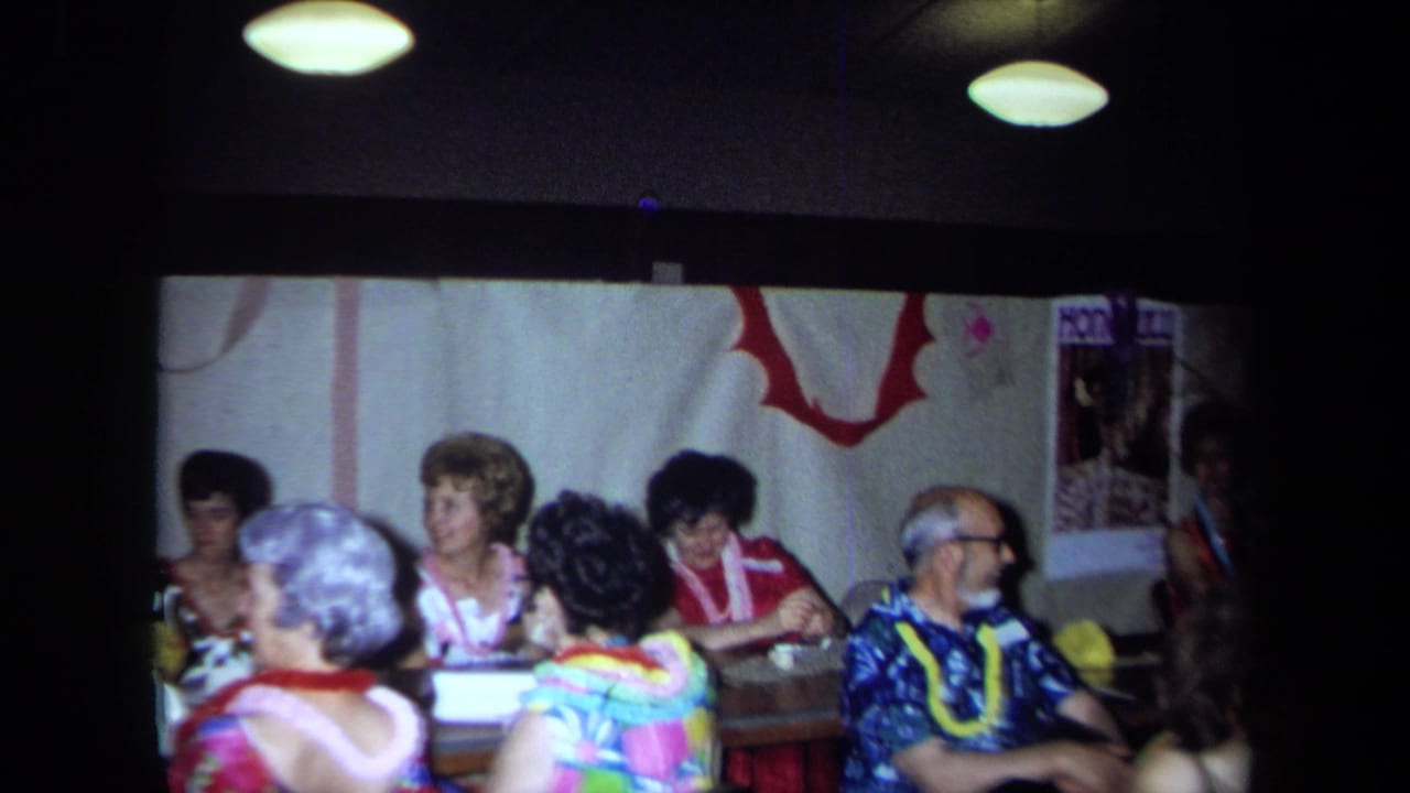 1971 California home-movie frame of adults at a Hawaiian-themed indoor party with leis, streamers, and table seating.