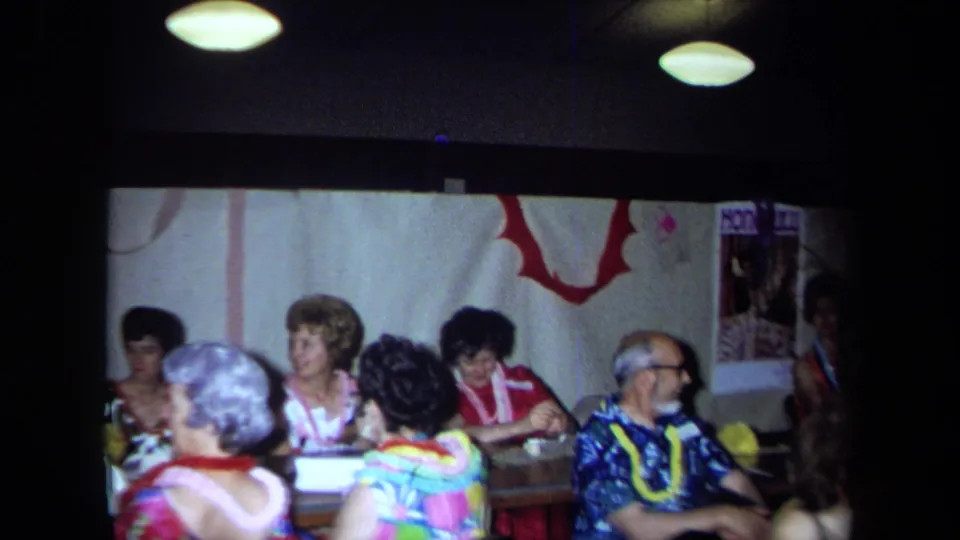 1971 California home-movie frame of adults at a Hawaiian-themed indoor party with leis, streamers, and table seating.
