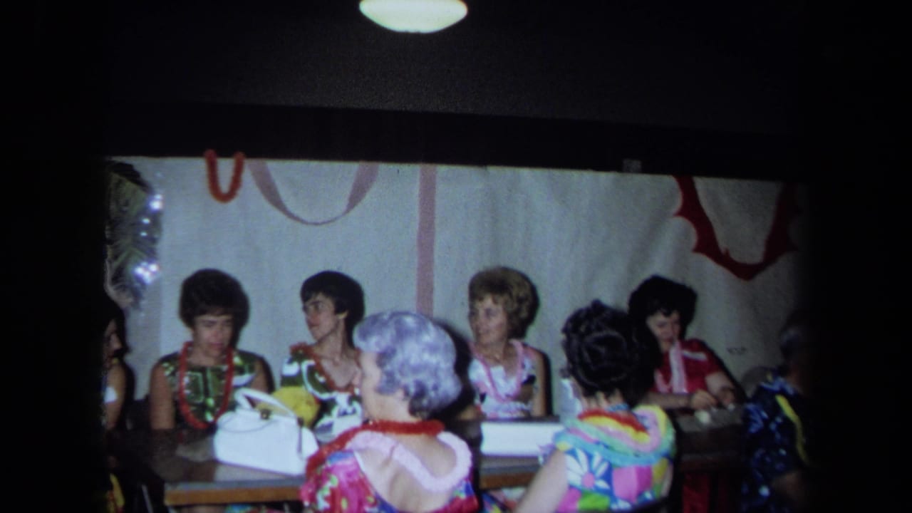 Party guests in leis sit around a table with a white handbag nearby.