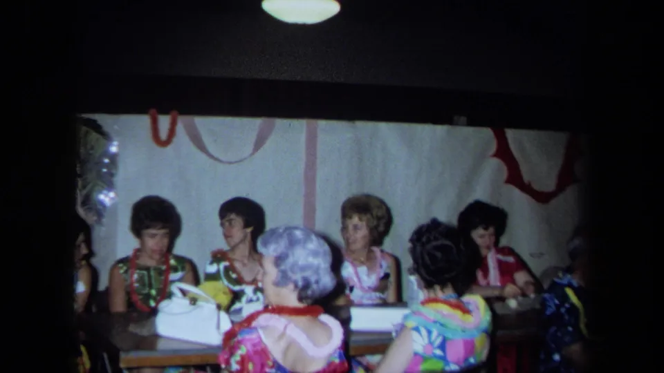 Adults at a 1971 California Hawaiian-themed party sit in conversation beside a white handbag and wall decorations.