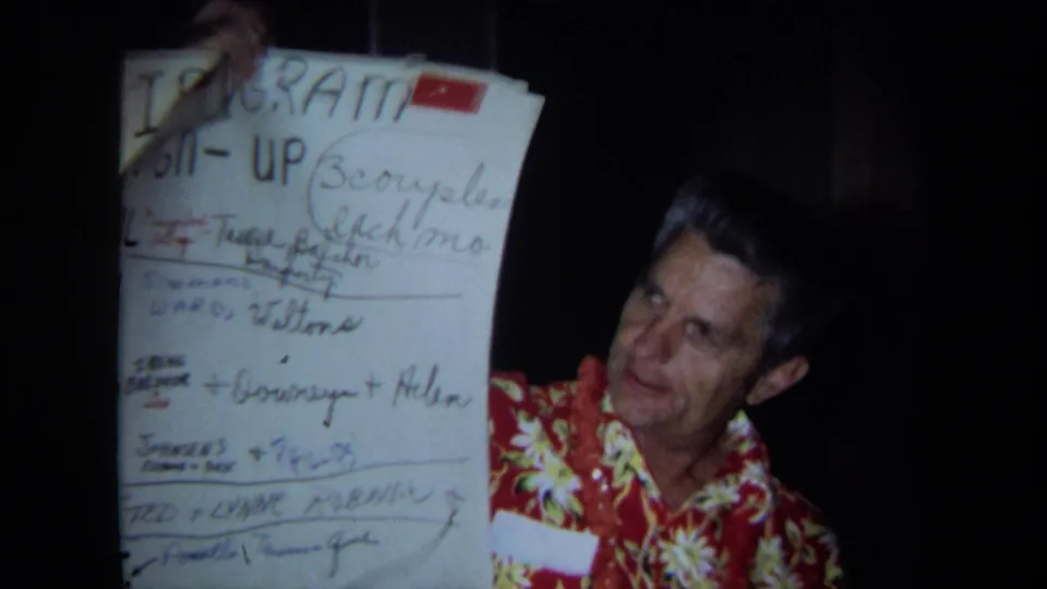 A man in an aloha shirt holds a handwritten party program in an indoor 1971 California home-movie scene.