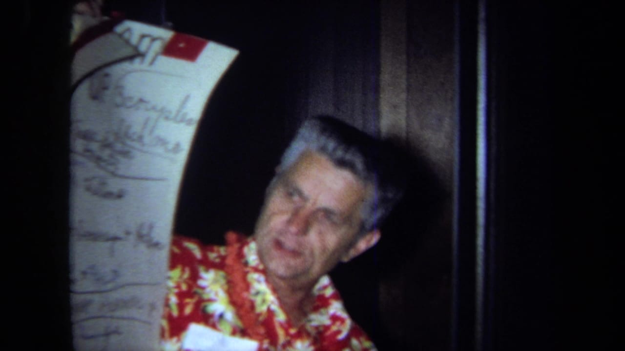 In 1971 California home-movie footage, an older man in a red floral shirt and lei holds a tall handwritten schedule chart indoors.