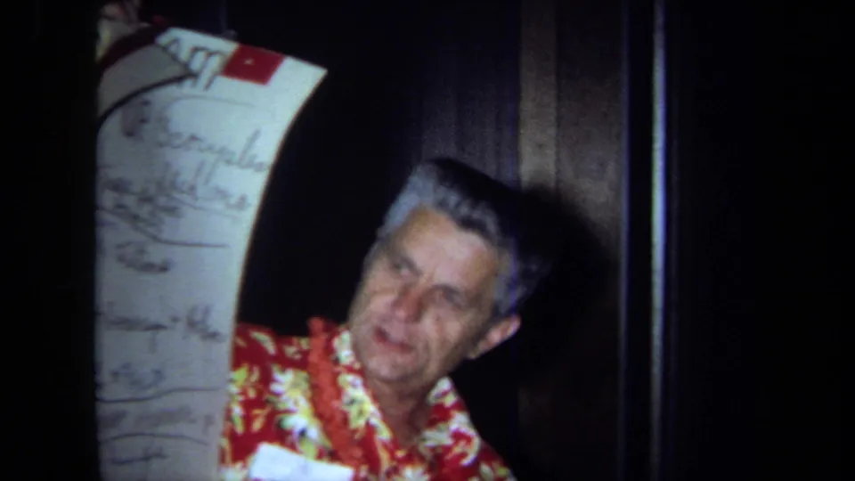 In 1971 California home-movie footage, an older man in a red floral shirt and lei holds a tall handwritten schedule chart indoors.