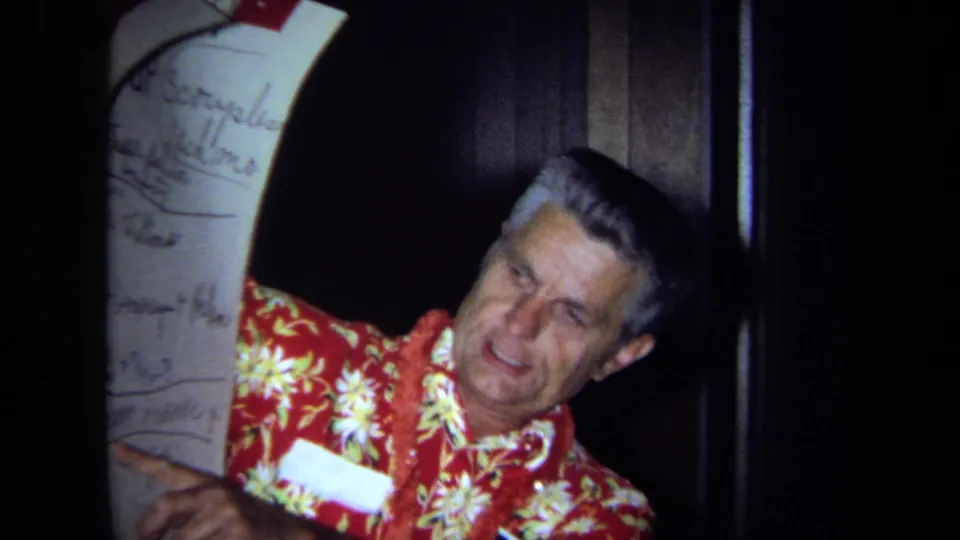 The presenter gestures beside a large handwritten monthly chart in a dim California interior, with 8mm grain and soft focus visible.