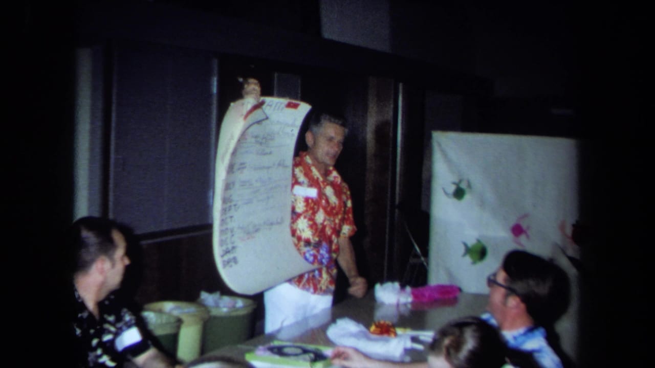 In a 1971 California home-movie frame, a man in a red floral shirt presents a large handwritten list to seated adults indoors.