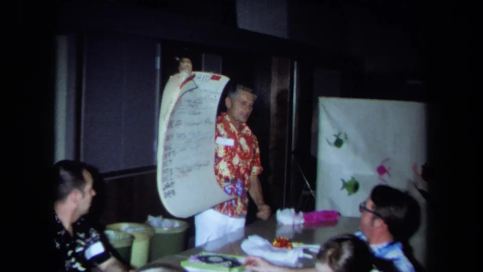 In a 1971 California home-movie frame, a man in a red floral shirt presents a large handwritten list to seated adults indoors.