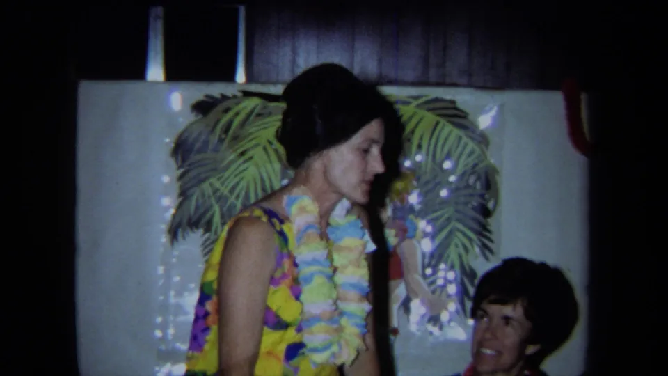 The home-movie frame shows a lei-wearing woman beside seated guests, with palm-frond decor and a hula dancer poster behind her.