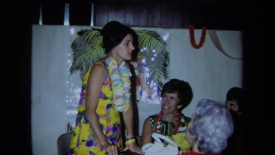 A woman in a tropical dress and leis stands near seated women in an indoor Hawaiian-themed 1971 California home-movie scene.