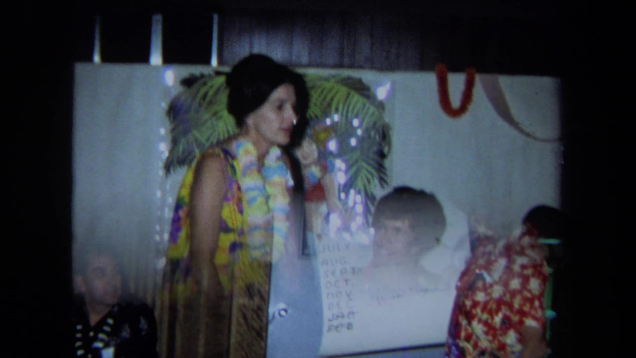 Woman in a floral dress and lei stands near a handwritten chart during an indoor party.