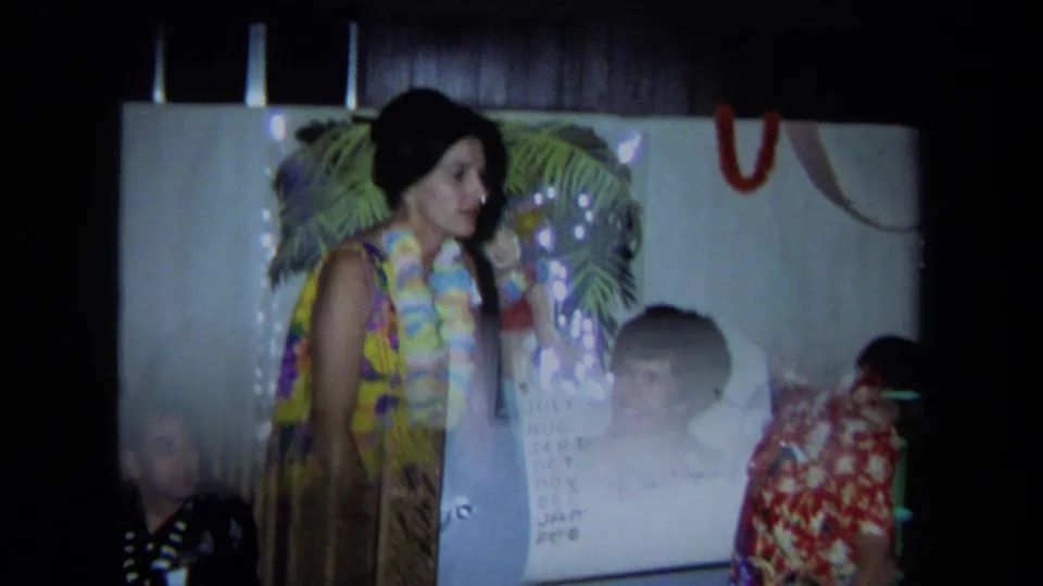 1971 California home-movie frame showing a woman in a floral dress and lei near a palm-frond backdrop while guests watch a chart presentation.