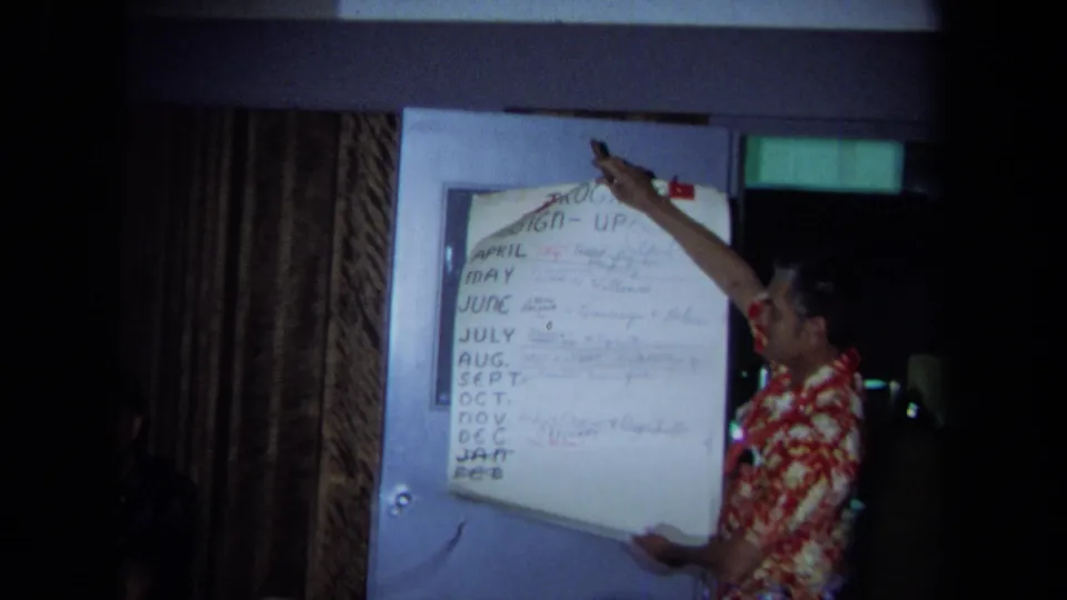 1971 California home-movie frame with a red aloha shirt presenter pointing at a large sign-up chart listing months in a private interior.