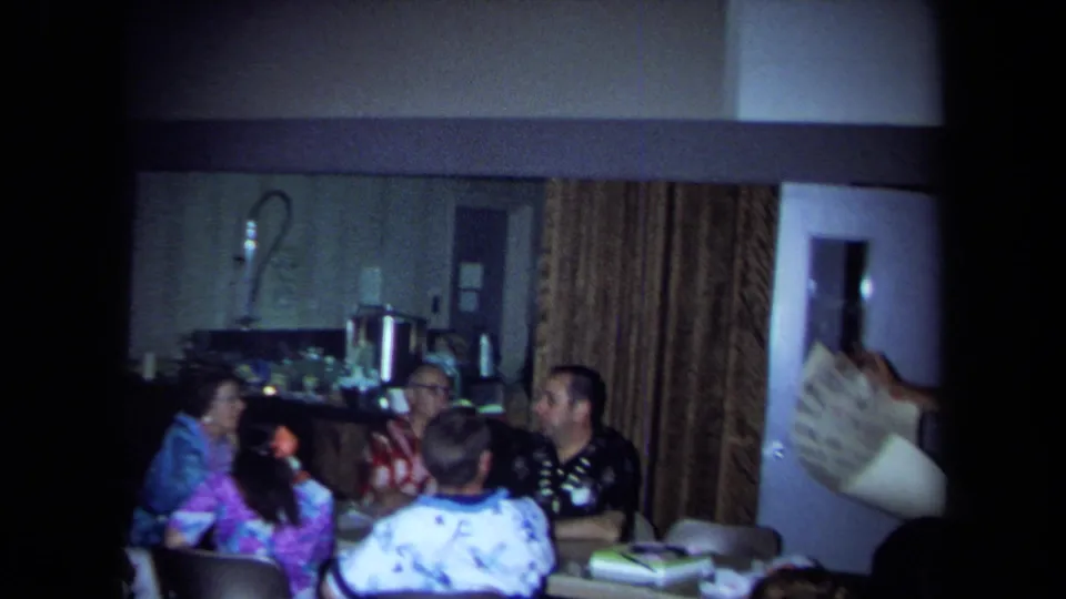 1971 California home-movie frame showing seated party guests watching or discussing a handwritten chart in a dim private room.