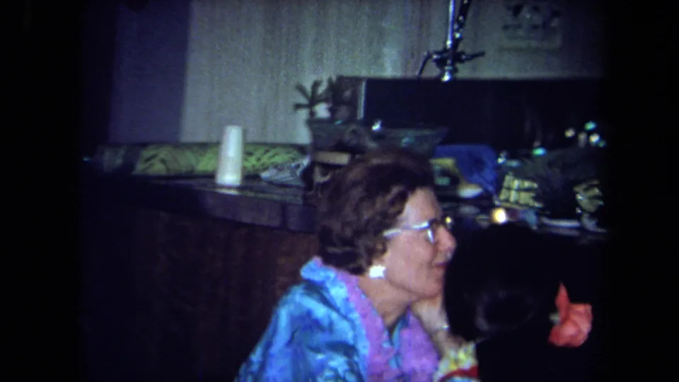1971 California home-movie frame showing a laughing woman in a blue party top beside a kitchen counter in a private interior.