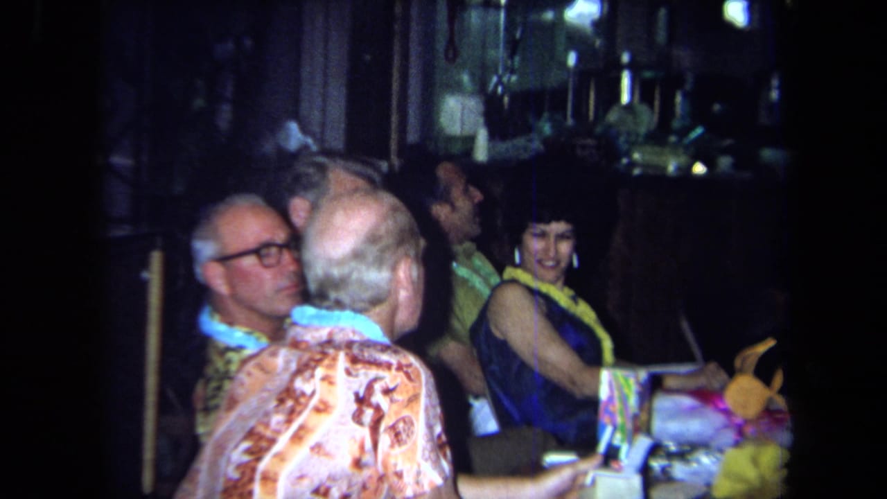 Adults in leis sit at a table with drinks and bright party decorations in a dim room.