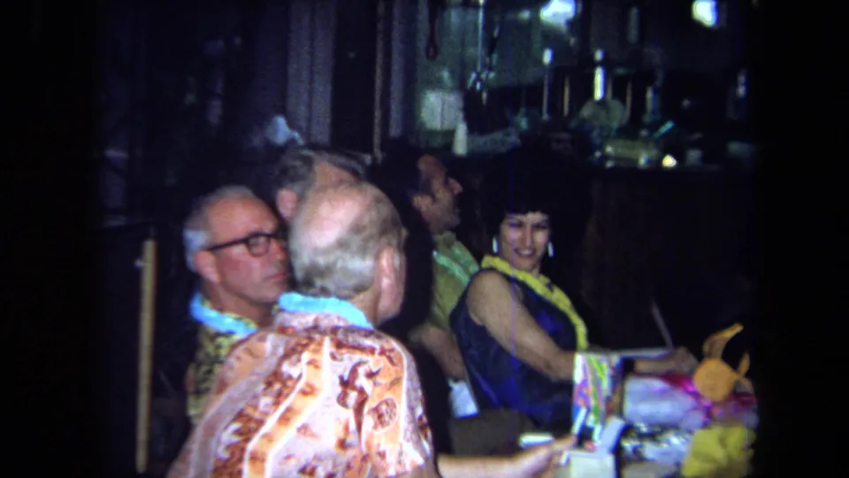 Adults wearing leis and aloha-style clothing gather around a table with drinks in a dim 1971 California party interior.