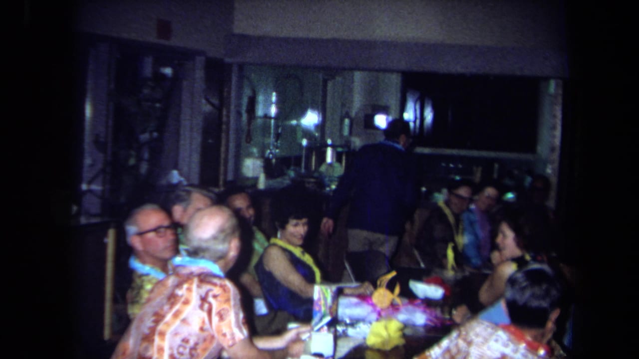 A group of adults in leis and patterned shirts sit around a festive table in a low-lit California interior from 1971.