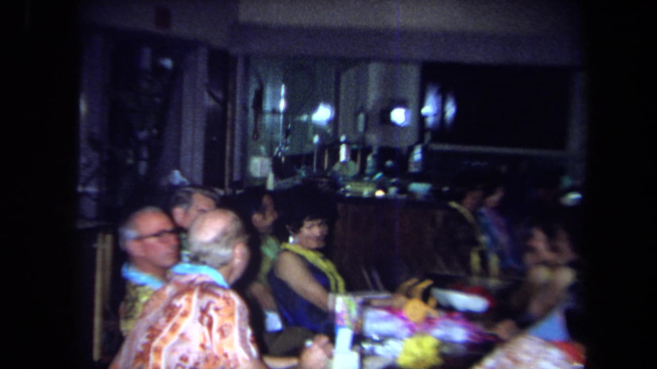 Adults wearing leis and colorful shirts talk at a party table, with a bar counter visible in the 1971 California home-movie frame.