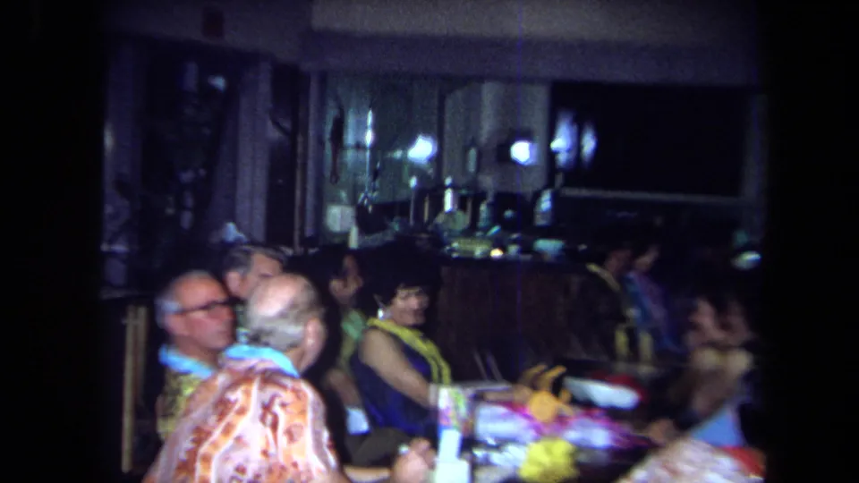 Adults wearing leis and colorful shirts talk at a party table, with a bar counter visible in the 1971 California home-movie frame.