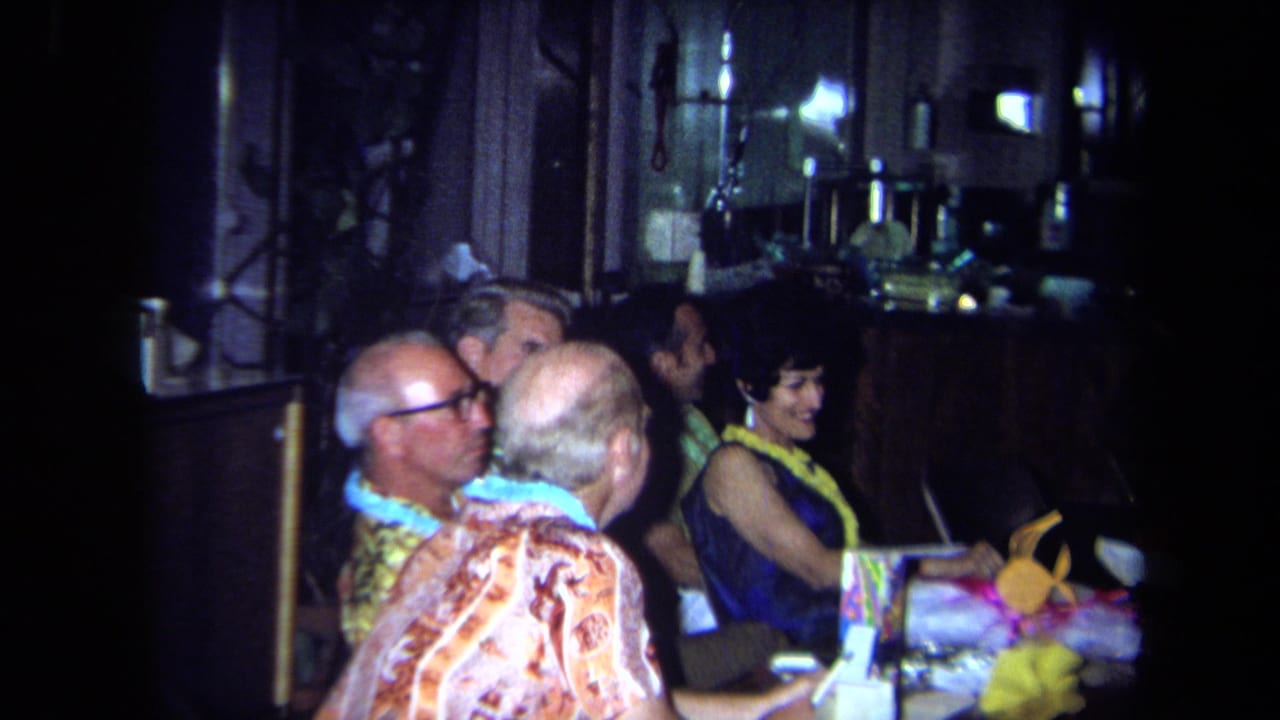 1971 California home-movie frame of older adults at a dim restaurant table during a Hawaiian-themed party.