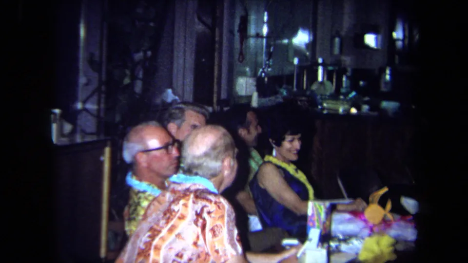 1971 California home-movie frame of older adults at a dim restaurant table during a Hawaiian-themed party.