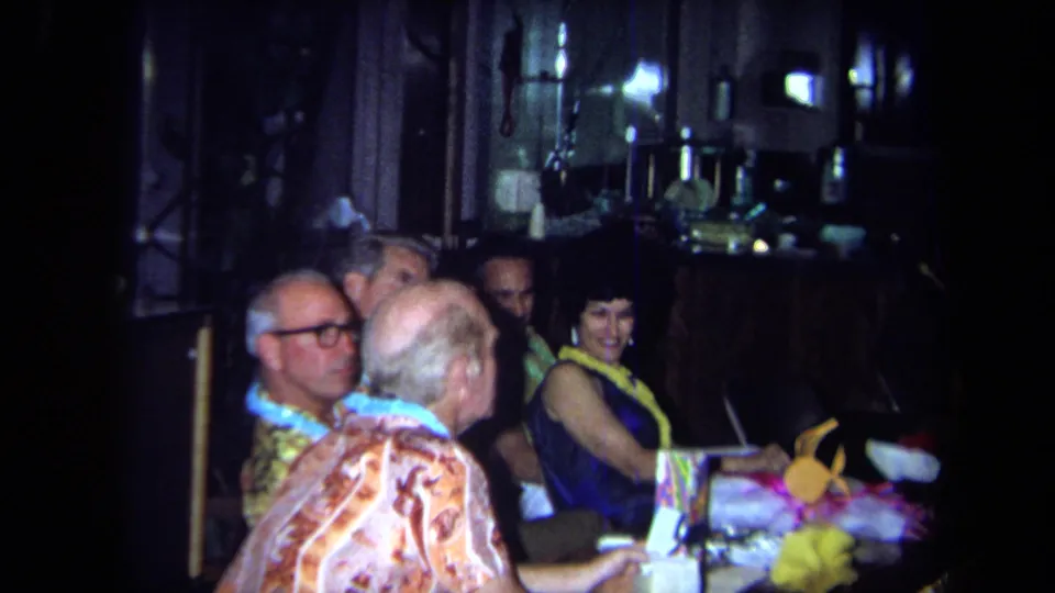 1971 California restaurant party frame with older guests in leis and a smiling woman in a blue dress at the table.