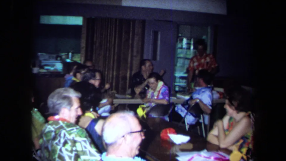 Wider 1971 California home-movie view of older adults seated in a restaurant during a Hawaiian-themed party.