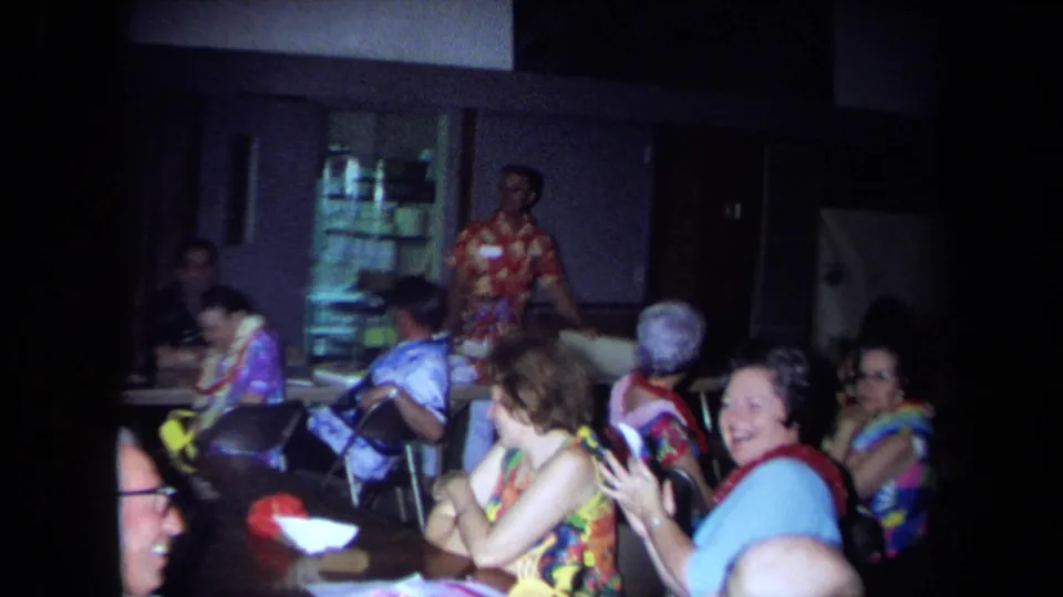 1971 California restaurant party frame with guests clapping, laughing, and a man in a red patterned shirt standing near the room.