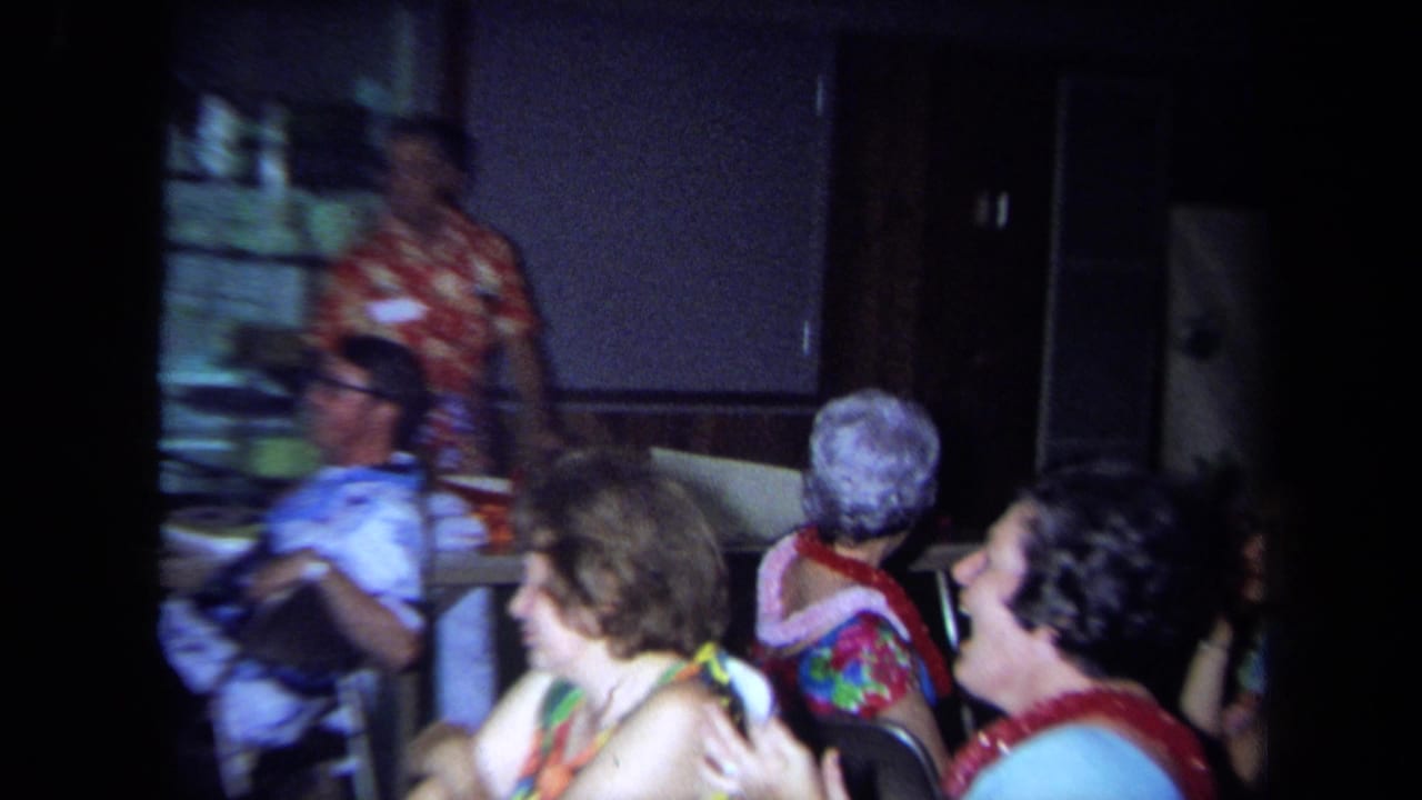 1971 California home-movie frame of women wearing leis seated at an indoor gathering as a man in a floral shirt stands at the front.