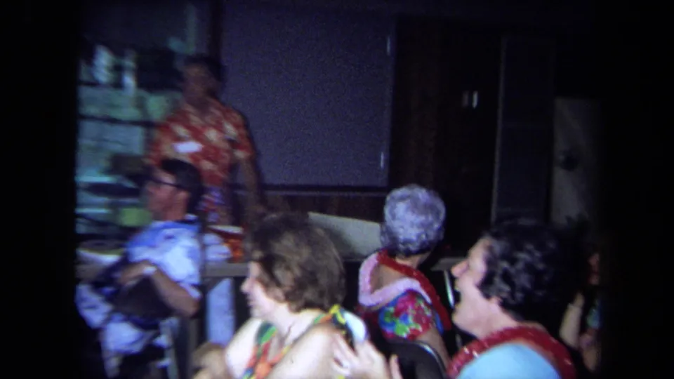 1971 California home-movie frame of women wearing leis seated at an indoor gathering as a man in a floral shirt stands at the front.