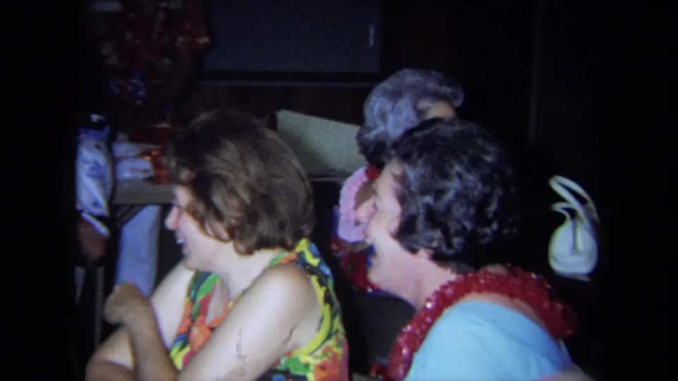 1971 California 8mm frame showing women in colorful tops and leis seated indoors during a casual social gathering.