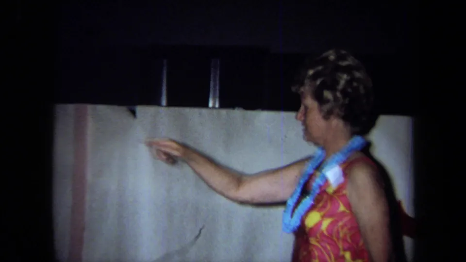 1971 California home-movie frame of a woman wearing a lei and sleeveless patterned top pointing inside a private room.
