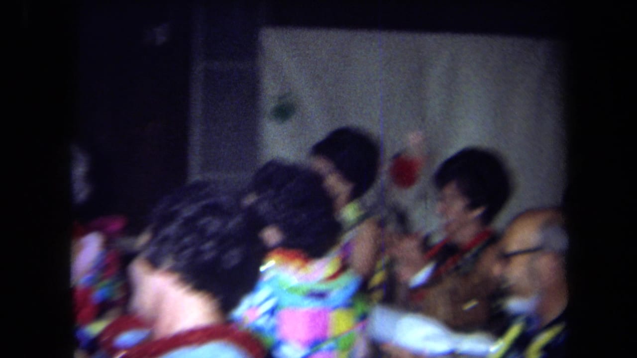 1971 California home-movie frame showing adults in colorful leis and patterned clothes seated close together during an indoor party.
